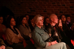 Koncert: Pracownia piosenki prezentuje - Niepozorny zaścianek - Zdjęcie pokazuje widownię w Piwnicach TCK podczas koncertu. Widzowie, siedzący na krzesłach obserwują występ. Na zdjęciu widać różnorodność publiczności, od młodych po starszych słuchaczy. Sala jest delikatnie oświetlona.