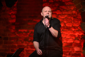 Koncert - Pracownia Piosenki "O jesieni..." - Tomasz Wiśniewski, aktor, występuje z mikrofonem na tle czerwono oświetlonej ściany.